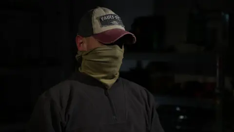 Kevin McGregor/BBC Ruslan, Special Operations Forces with a hat and snood hiding his face and identity in a dark room wearing a black fleece