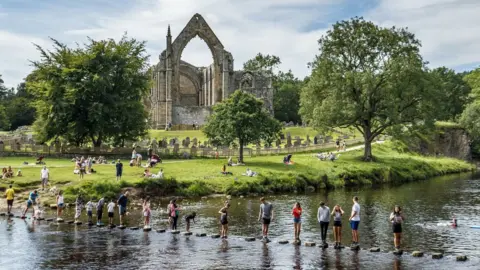 PA Media Bolton Priory overlooks the River Wharfe as families cross on stepping stones