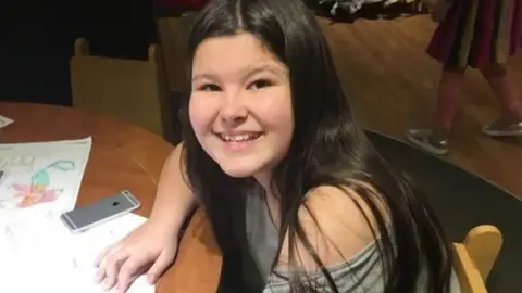 Jayne Reynolds A teenage girl smiles at the camera while sat down leaning on a table. She has long, dark hair. 