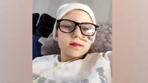 Sophie Robinson - a young girl with a soft white hat on - is wearing large black-rimmed glasses and has a tube coming out of her nose with a sticker on it. She is wearing pink lipstick and a sequined white top and looking down into the camera.