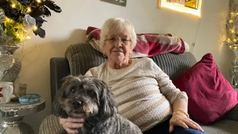 An elderly woman with short, blond hair and wearing glasses and a striped cream and brown jumper holding a pet dog which has long ears and brown/grey whiskers while they sit on a sofa (He tried to bite me, playfully!)