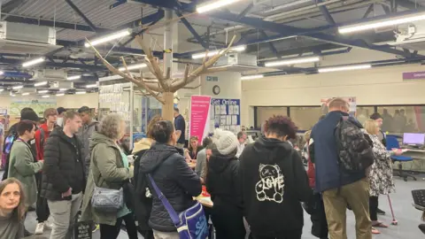 West Northamptonshire Council A crowd of people around a desk waiting to speak to a hub helper. 