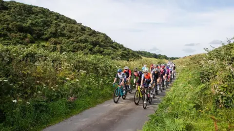 Gran Fondo Isle of Man A crowd of cyclists in brightly coloured jerseys and helmets on bicycles travel on a tarmac road with greenery on either side.