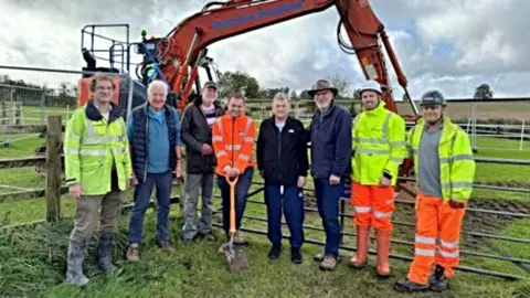 East Devon District Council A group of eight councillors and contractors are photographed in a field with an orange digger behind them. Those photographed are seen in high viz clothing.