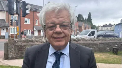 A man with grey hair and rectangular glasses smiles for a head and shoulders shot. He is wearing a dark grey suit with a light blue shirt and dark blue tie.