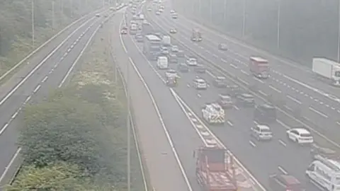 A traffic cam showing a motorway at a junction. The road is lined with thick woodland. On the left, a slip road joins the main motorway which is heavy with traffic moving away from the camera. The other side of the central reservation contains lighter traffic and a white lorry is leaving via a slip road which disappears from view.