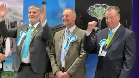Three men are wearing turquoise ties, grey suits and blue and white Reform rosettes. They are stood in a line clapping and punching the air in celebration.