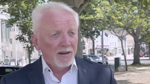 BBC Lee Madden is wearing a black blazer and white shirt, he has white hair an blue eyes. 