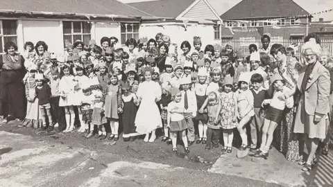 A black and white picture of a large group of children of more than 40 children with at least 10 adults among them, The children are party outfits, including dresses and skirts, while one of the boys wears a tie. Some of the children are wearing crown hats. 