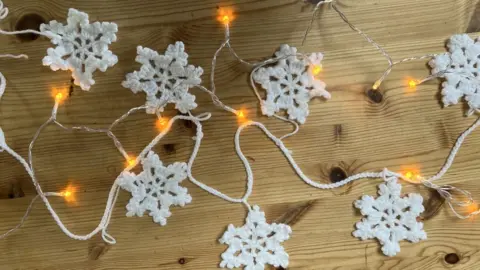 Marta Pulencika a crocheted snowflake garland on a wooden table alongside fairy lights