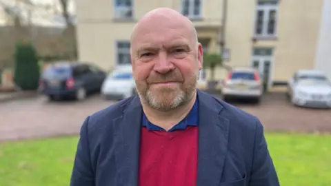 Council leader Bill Revans looking at the camera while wearing a red jumper and grey blazer