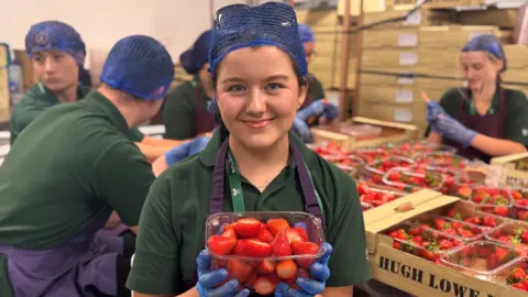 Emily Ostrowska wearing a green uniform with a blue hairnet and gloves, smiling for the camera. She is holding a punnet of strawberries with the green stalks removed. Six other people in identical uniforms work on trays of berries on a table in the background.