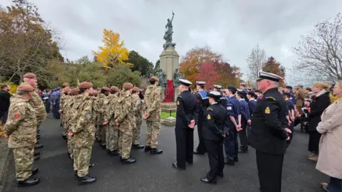 Johnny Rutherford/BBC Personnel from the armed forces gathered around a cenotaph in Exeter. They have their hands behind their back. Trees in autumnal colours are seen in the background on a cloudy day.