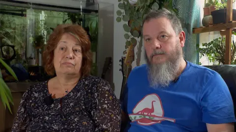BBC Amanda Hoyes sitting on a sofa next to her partner Chris Davis. Amanda, with shoulder length brown hair, is on the left wearing a dark patterned round neck top and Chris, with short hair, a beard and moustache, is wearing a blue t-shirt.