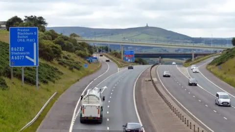 Geograph/Mat Fascione Vehicles on either side of the M65 at a slip road junction, with Darwen Hill and the Jubilee Tower on the summit in the background