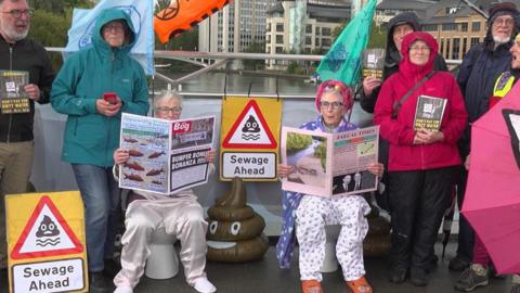 A group of protesters sitting on a bridge over the River Thames. Two are sitting on toilets while six are standing and holding banners