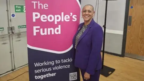 West Yorkshire Combined Authority A woman with a shaved head and wearing a purple suit and black and cream checked blouse smiles at the camera as she poses next to a large sign which says The People's Fund in white capital letters on a pink background above the words Working to tackle serious violence together in white letters on a dark blue background.