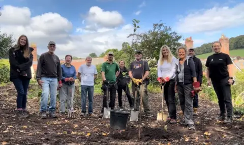 NHS Somerset ICB A group of 12 people are standing in a muddy field, with many of them holding shovels and forks. They are all looking at the camera, smiling.