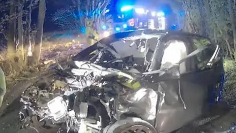 Lancashire Police Mangled car wreckage lies on the ground among trees. The front of the grey Audi is completely smashed.