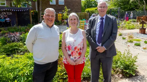 Telford and Wrekin Council Two men and a woman stand in front of a garden featuring plants, pathways and children's furniture.