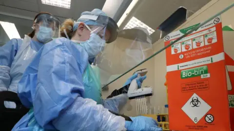 Two medical technicians are wearing PPE at Queen Elizabeth University Hospital in Glasgow on April 22, 2020. Both are wearing blue, plastic aprons, surgical masks and visors. There is a sign behind them which says "bio-bin" and appears to be a place to deposit used medical equipment and PPE.