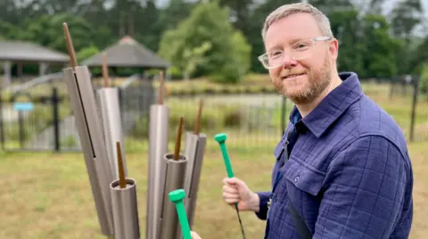 Stuart Wood stands next to a set of tubular bells with a pond behind him while holding two chime hammers.