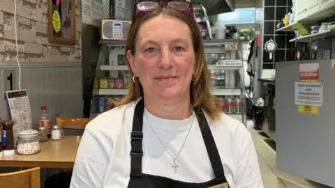 Helen Lawson has shoulder length brown hair and a white T-shirt with a black apron over it. 