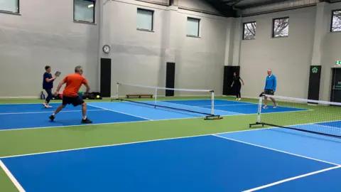 BBC/Julie Mariotti Four people are playing pickleball in The Pickleball Shed, which is a large warehouse space with green and blue flooring and white nets. The players are dressed in sports wear and are each holding a black pickleball racket.