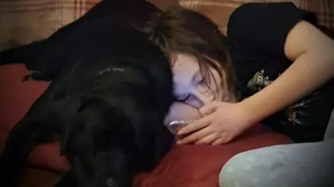 Elizabeth Kane A teenage girl lies on a red sofa, beside her is a black Labrador