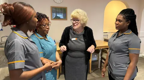 Steve Hubbard/BBC Sue Watson with three care workers. All are standing in a room with a table behind them and appear to be listening to watson speak. 
