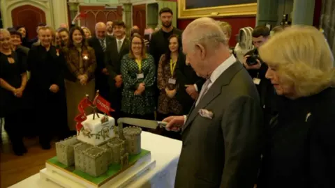 King Charles cutting a cake in the shape of a castle, surrounded by people watching