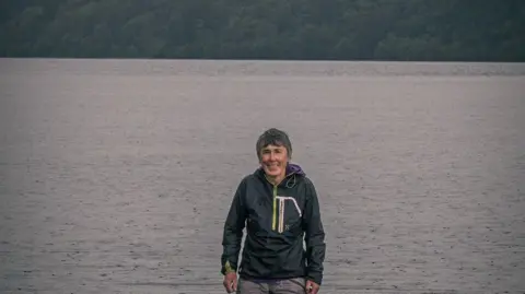 Amy Bateman A woman with cropped grey hair standing in front of a lake.