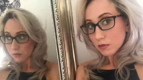 A woman with blonde hair and dark rimmed glasses stands next to a gold rimmed mirror which shows her full reflection