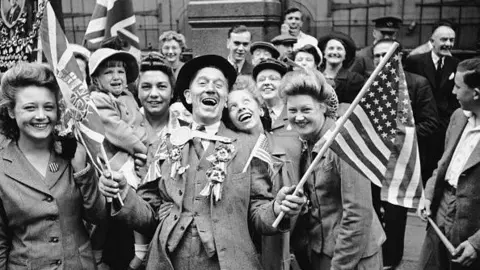 Getty Images Crowds celebrate VE Day at the end of World War Two on 8 May 1945. A man in a suit and hat is holding flags in each hand and leaning back and laughing, and women are all around him looking happy. One of the women is holding a crying child. There are more people in the background in the black and white archive photograph.