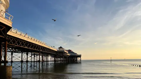 Owen Sennitt/BBC The view of a Victorian pier with metal structures holding it up from the beach as seagulls fly above on a sunny morning.