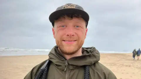 Wearing a dark green coat with a black dog lead hanging round his neck, dog walker Dan smiles at the camera. He also wears a grey baseball cap, with a light ginger beard and blue eyes. 