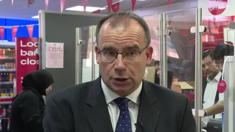 Post Office Minister Gareth Thomas in suit and tie, standing in front of a Post Office counter, with customer and staff in the background