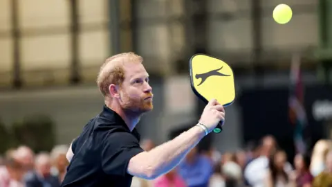 A man in a black shirt with red hair and beard holding a pickleball bat with a yellow cover and black cat image. He is about to hit a yellow ball