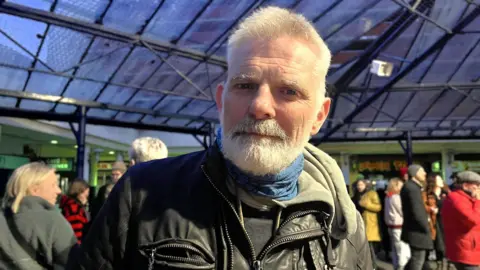 Maddy Jennings/BBC Simon Floyd stood in front of dozens of people. He is wearing a black leather jacket. He has short grey hair and a grey beard