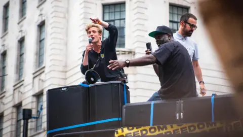 Save Our Scene George Fleming speaking into a microphone standing on a sound system outside