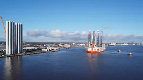 RWE An image of the Wind Peak as it sails down the Humber Estuary towards Hull. The large, red and white vessel is flanked by two red and white tug boats. A passenger ferry is visible in the distance. The vessel is carrying four tall black towers.