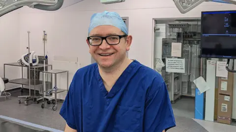 University of Leicester Matt Bown smiling wearing black-framed glasses and blue medical scrubs.