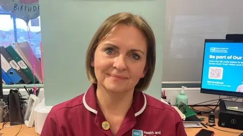 Carol is looking into the camera and smiling. She has chin-length brown hair and is dressed in maroon medical scrubs. She is pictured in a clinical environment, behind her is a busy desk with a number of lever arch files, documents and a computer screen.