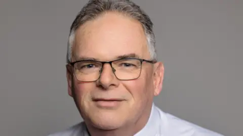 Colin Lang, a man who is looking directly at the camera and smiling. He is wearing a white shirt and red tie.