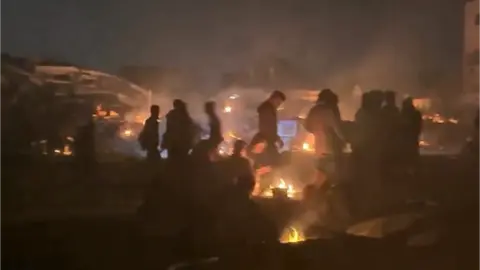 Instagram Still from an Instagram video showing figures waiting next to campfires in the dark.