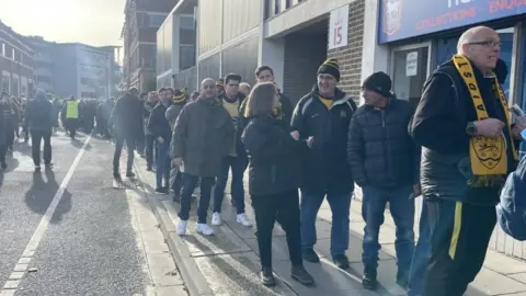 A queue of men and women wearing Maidstone United scarves outside the Ipswich Town Stadium