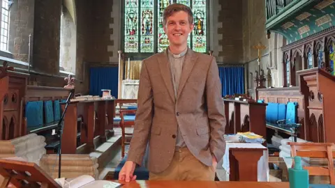 Tom Houston Man with short light brown hair and brown jacket with clerical collar stands in a church