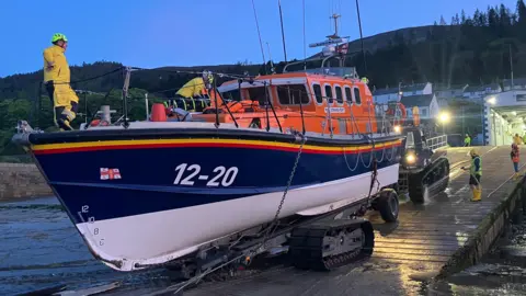 BBC An orange, blue and white boat that is docked in the evening on a ramp with men in yellow standing on the boat and deck and fixing maintaining it.