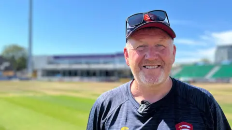 A man wearing sunglasses and a sun hat at the top of his head while standing on the cricket pitch. He has a beard and he is smiling at the camera. 