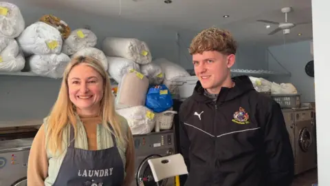 A woman with long blonde hair, wears a navy apron which says 'laundry' on and stands in a laundrette next to her son, who is tall, with curly blonde, short hair, wearing a black coat with the Altrincham FC emblem on 